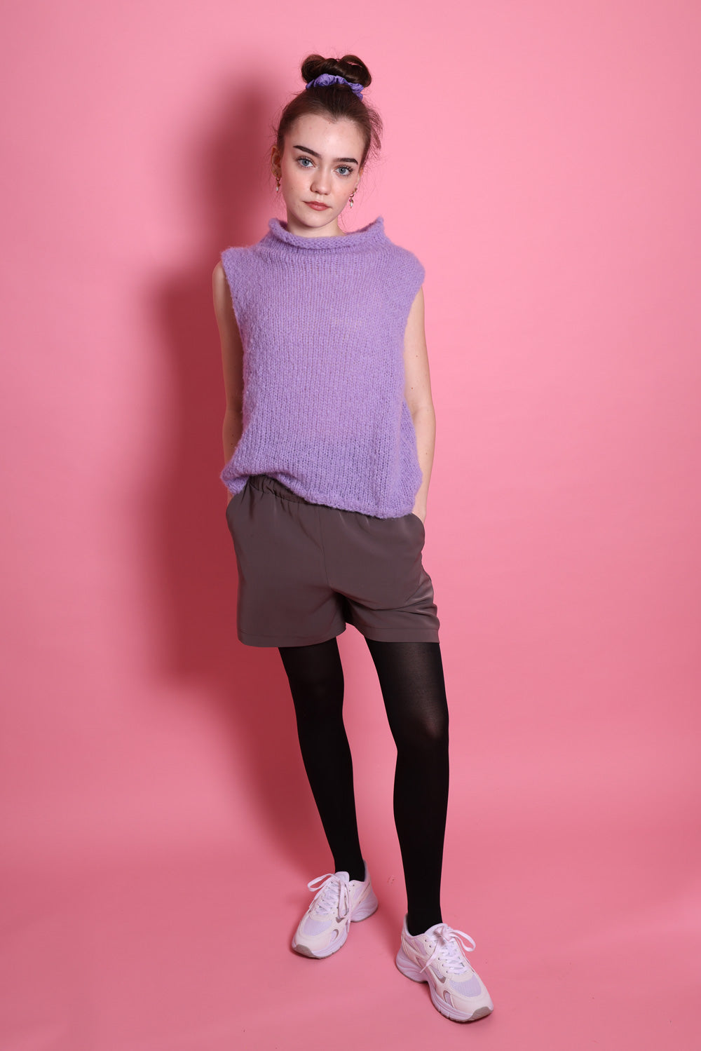 Ein Junges Teeniegirl mit dunklen, zum Scrunchie und einem lilafarbenem Wollpullunder steht vor einer rosa Wand. Sie ist ein Senior. Sie trägt eine graue Short und eine schwarze Strumpfhose und weiße Schuhe. Ihre Hände sind in den Hosentaschen. Sie steht mit frontal zum Betrachter.
