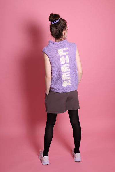 Ein Junges Teeniegirl mit dunklen, zum Scrunchie und einem lilafarbenem Wollpullunder steht vor einer rosa Wand. Sie ist ein Senior. Auf der Rückseite des Pullunders ist das Wort Cheer eingestrickt. Die Buchstaben laufen untereinander und sind weiß. Sie trägt eine graue Short und eine schwarze Strumpfhose und weiße Schuhe. Ihre Hände sind in den Hosentaschen. Sie steht mit dem Rücken zum Betrachter.