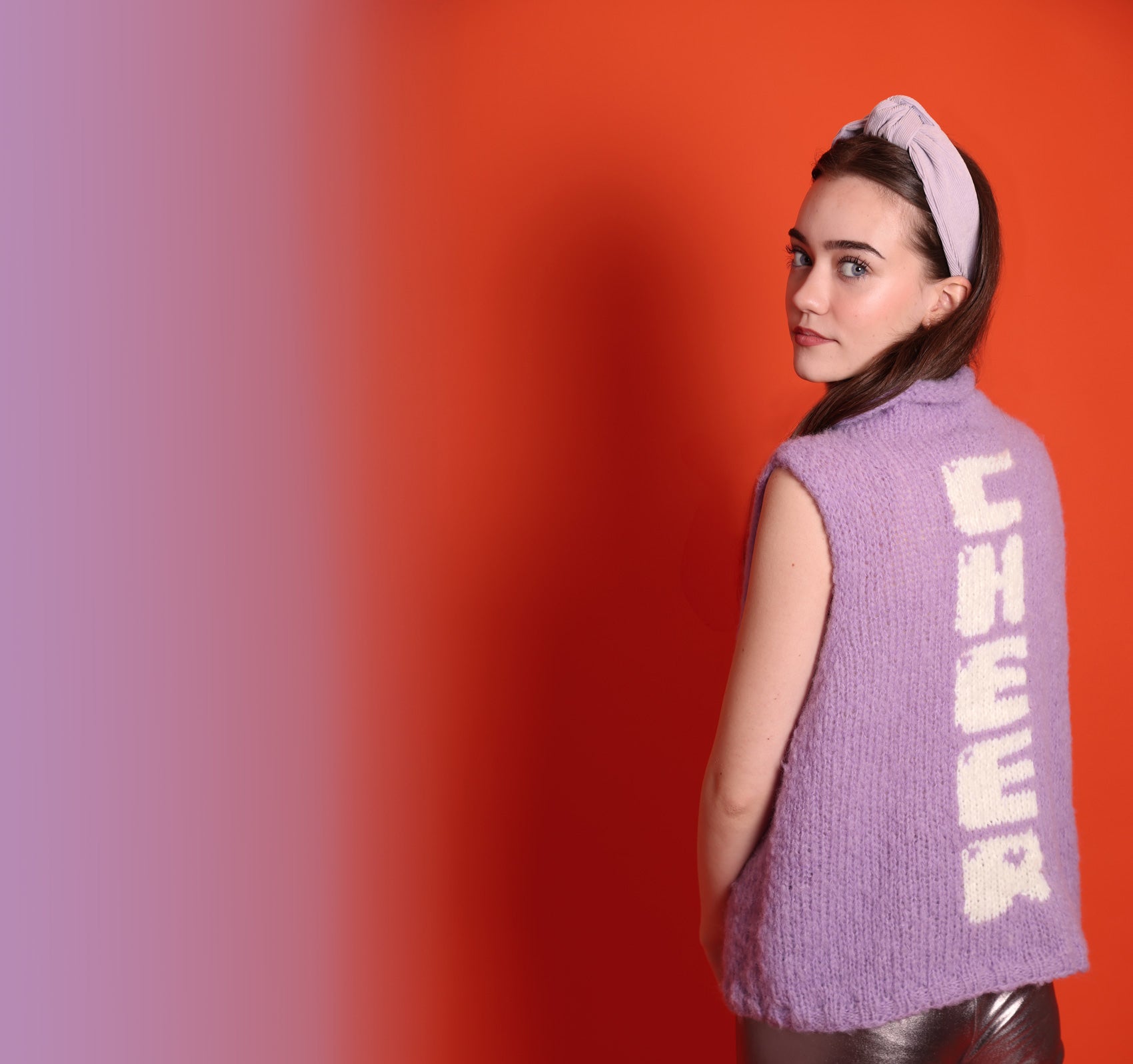 Ein junges Teeniegirl mit dunklen offenen Haaren, einem lila Haarreif und einem lilafarbenem Wollpullunder steht vor einer orangefarbenen Wand. Rechts ist ein Verlauf in lila. Sie trägt zudem eine silberne Leggings. Sie ist ein Senior. Auf der Rückseite des Pullunders ist das Wort Cheer eingestrickt. Die Buchstaben laufen untereinander und sind weiß. 