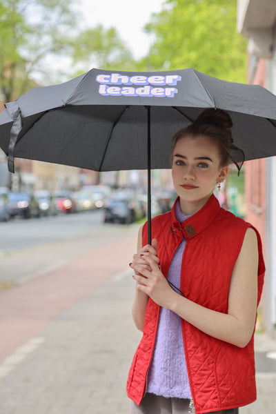 Mädchen mit roter Weste und lila Pullunder, dunklen Haaren im Dutt hält draußen einen schwarzen Regenschirm mit der Aufschrift: "CHEERLEADER" in Pixelart..