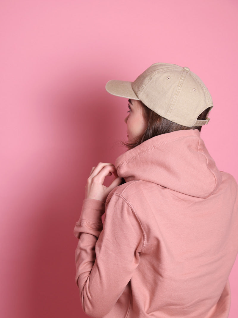 Person von hinten mit rosafarbenem Pullober vor rosafarbenem Hintergrund mit dunklen Haaren und beiger Cap.