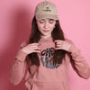 junges Teenie-Mädchen trägt einen altrosafarbenen Pullover mit Aufschriff Cheerleader und Kangaroopocket, eine graue Hose, roten Nagellack, offene dunkle Haare und eine beige Cap mit "I BOW Cheer" drauf. Die Schleife hat eine leichte Herzform. 