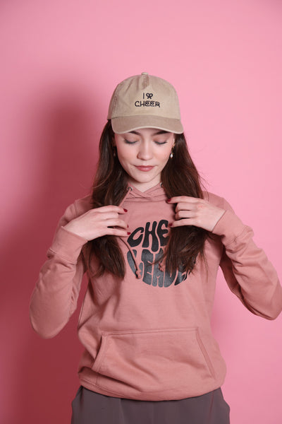 junges Teenie-Mädchen trägt einen altrosafarbenen Pullover mit Aufschriff Cheerleader und Kangaroopocket, eine graue Hose, roten Nagellack, offene dunkle Haare und eine beige Cap mit "I BOW Cheer" drauf. Die Schleife hat eine leichte Herzform. 