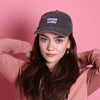 Mädchen mit langen dunklen Haaren und blauen Augen trägt eine graue Vintage releaxed Cap in grau mit lilafarbenem Aufdruck: "Cheerleader" in PixelArt. Sie trägt einen rosa farbenen Pullover und steht vor einer rosafarbenen Wand.