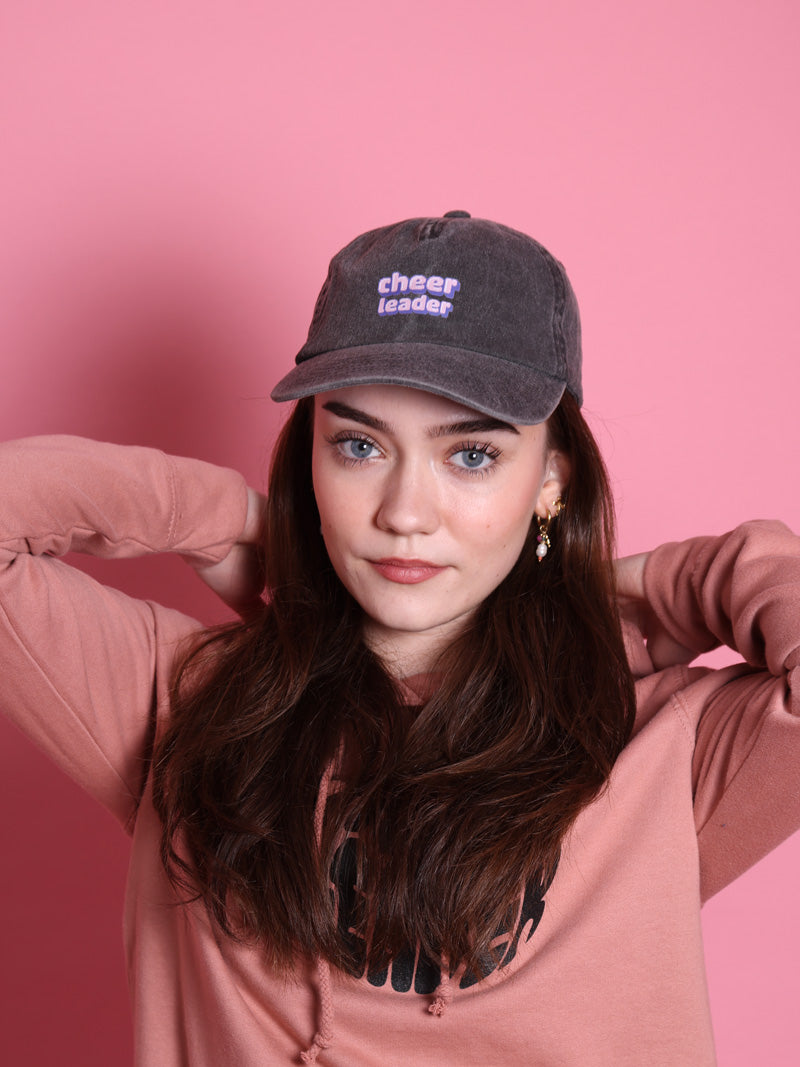 Mädchen mit langen dunklen Haaren und blauen Augen trägt eine graue Vintage releaxed Cap in grau mit lilafarbenem Aufdruck: "Cheerleader" in PixelArt. Sie trägt einen rosa farbenen Pullover und steht vor einer rosafarbenen Wand.