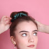 Teeniemädel mit dunklen Haaren und grünem Satinscrunchie steht vor rosa farbener Wand. Sie hat Perlen- und Schleifenohringe drin, blaue Augen und helle Haut. Sie trätgt etwas mit lilafarbenem Wollkragen.