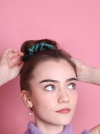 Teeniemädel mit dunklen Haaren und grünem Satinscrunchie steht vor rosa farbener Wand. Sie hat Perlen- und Schleifenohringe drin, blaue Augen und helle Haut. Sie trätgt etwas mit lilafarbenem Wollkragen.