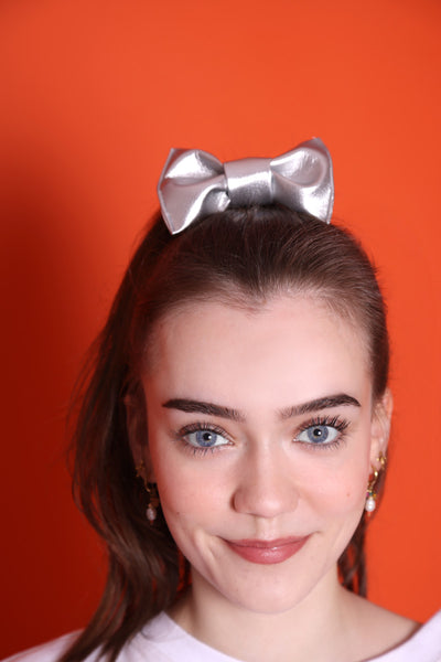 silberne Haarschleife aus Kunstleder sitzt auf einem Kopf eines Teenie-Mädchens, welches dunkle Haare, blaue Augen hat und Perlenohrringe trägt. Sie steht vor einem orangefarbene Hintergrund. 