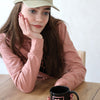 Mädchen sitzt an einem Holztisch vor einer weißen Wand. Sie trägt eine beige Cheer Capi, einen rosapullover und hat braune lange Haare und blaue Augen. Sie hält in ihrer Hand eine schwarze Tasse mir rosa Aufschrift: Best Cheer Friends.