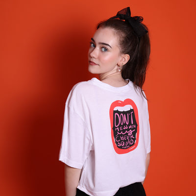 Zu sehen ist ein Senior Mädchen mit einem weißen Shirt mit dem Motiv: Don't mess with my cheer squad please. Dies steht in einem offenem Mund mit roten Lippen. Es handelt sich um eine Illustration. Sie steht mit dem Rücken zum Betrachter, schaut aber nach hinten. Sie hat blaue Augen und ein zartes Gesicht. Das Motiv ist auch ein Backprint. Sie hat dunkle Haare im Zopf mit einer schwarzen Haarschleife. Sie trägt Perlenohrringe und steht vor einer orangefarbenen Wand. 