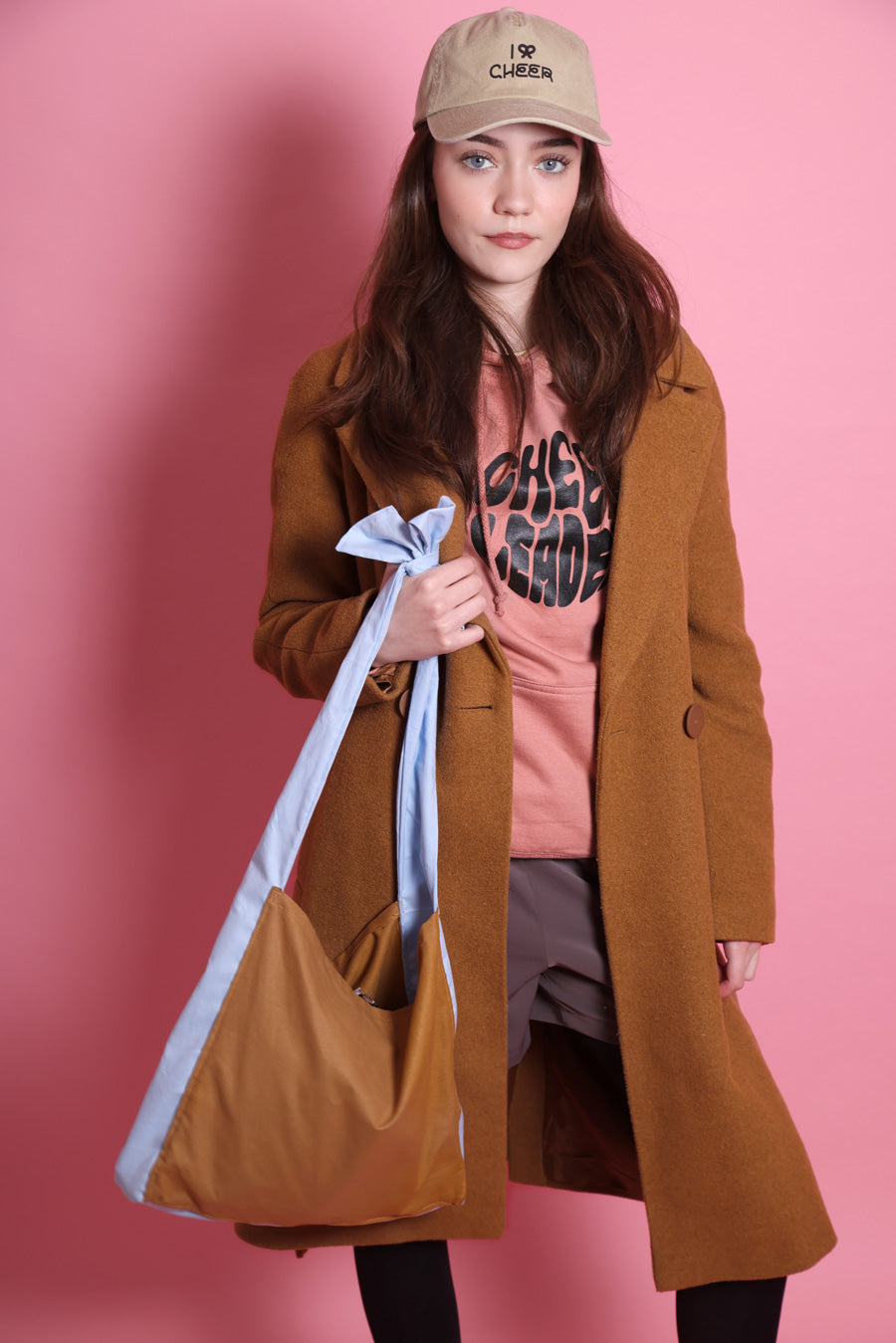 Shopper mit Schleife in karamel und hellblau wird von einem jungen Mädchen mit dunklen Haaren und blauen Augen festgehalten. Sie trägt eine Short mit schwarzer Strumpfhose, einen rosafarbenen Hoodie mit dem Aufdruck Cheerleader und eine beige Cap mit I Bow Cheer drauf. Sie steht vor rosafarbenem Hintergrund.