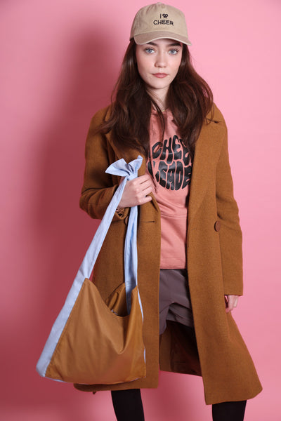 Shopper mit Schleife in karamel und hellblau wird von einem jungen Mädchen mit dunklen Haaren und blauen Augen festgehalten. Sie trägt eine Short mit schwarzer Strumpfhose, einen rosafarbenen Hoodie mit dem Aufdruck Cheerleader und eine beige Cap mit I Bow Cheer drauf. Sie steht vor rosafarbenem Hintergrund.