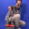 Mädchen mit silbernem Shirt trägt silber, graue-corallefarbene Tasche beige Hose, weiße Brille in Katzenform und weiße Schuhe. Sie steht vor einem blauen Hintergrund. 