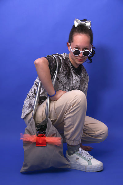 Mädchen mit silbernem Shirt trägt silber, graue-corallefarbene Tasche beige Hose, weiße Brille in Katzenform und weiße Schuhe. Sie steht vor einem blauen Hintergrund. 