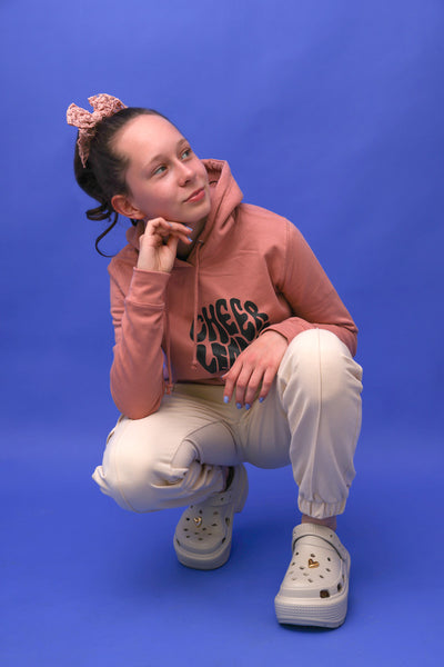 junges Teenie-Mädel mit rosafarbenem Hoodie, beiger Hose und Gummilatschen. Sie trägt eine rosa farbene Schleife aus Spitze und steht vor einem blauen Hintergrund. 