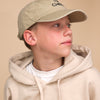 Junge mit beigem Pullover, beiger Cap steht vor beigem Hintergrund und schaut zur Seite. Auf seiner Cap kann man den schwarzen Schriftzug: "I BOW (symbolisch als Schleife) Cheer" nur erahnen.