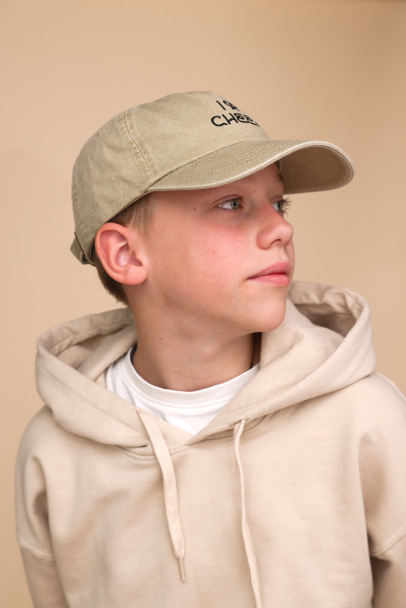 Junge mit beigem Pullover, beiger Cap steht vor beigem Hintergrund und schaut zur Seite. Auf seiner Cap kann man den schwarzen Schriftzug: "I BOW (symbolisch als Schleife) Cheer" nur erahnen.