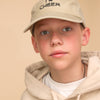 Junge mit beigem Pullover, herrgrünen Augen und beiger Cap mit schwarzer Aufschrift: I Bow Cheer steht vor einem beigem Hintergrund. 