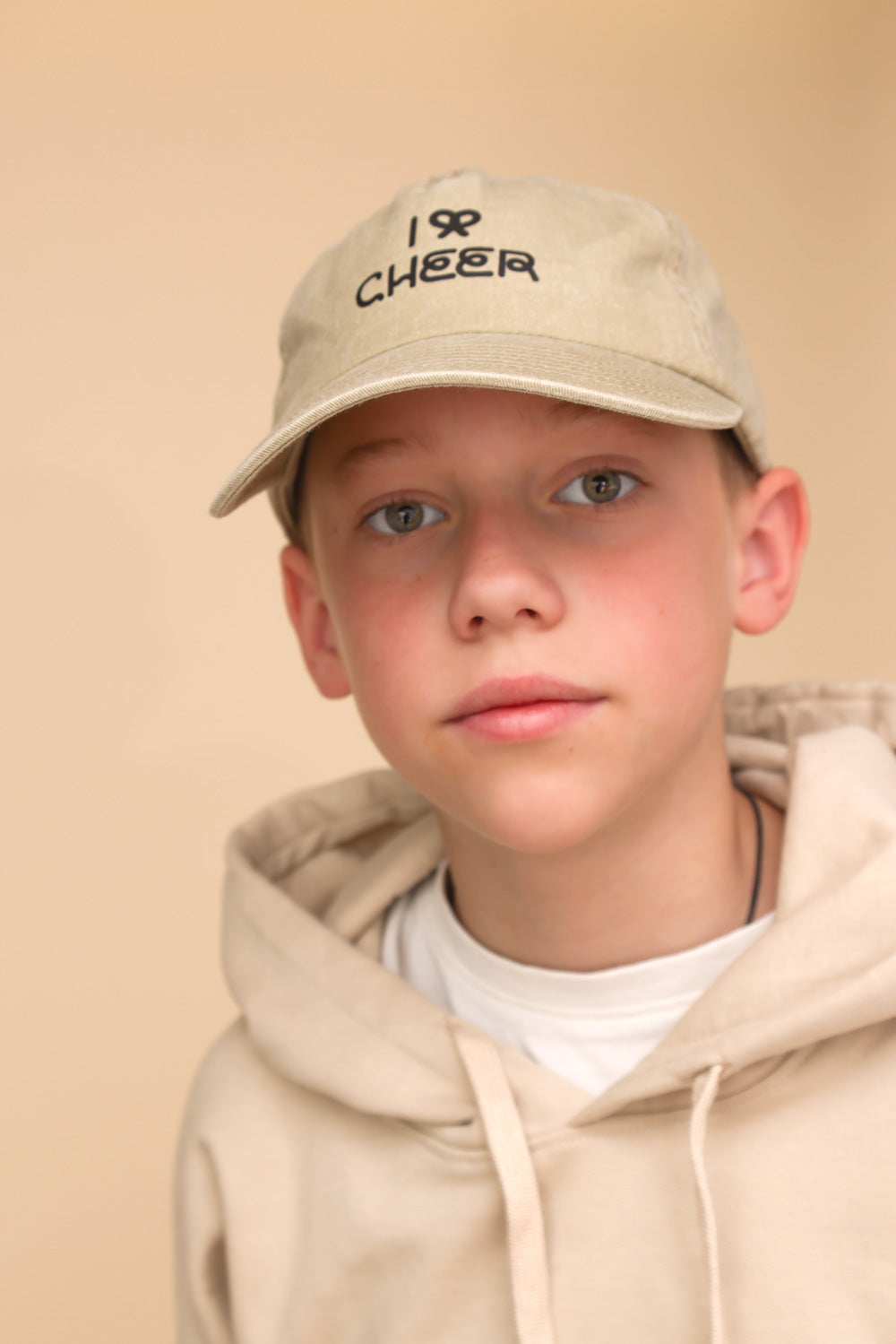 Junge mit beigem Pullover, herrgrünen Augen und beiger Cap mit schwarzer Aufschrift: I Bow Cheer steht vor einem beigem Hintergrund. 