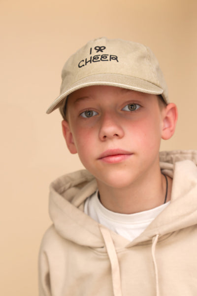 Junge mit beigem Pullover, herrgrünen Augen und beiger Cap mit schwarzer Aufschrift: I Bow Cheer steht vor einem beigem Hintergrund. 