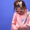 ein kleines Mädchen steht vor blauem Hintergrund. Sie trägt eine rosa Herzbrille, einen rosafarbenen Pullover mit dem Aufdruck: Cheerleader, eine Tasche mit silbernem Henke und eine bunt bestickte offwhie schleife.