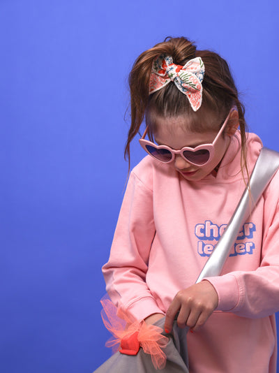 ein kleines Mädchen steht vor blauem Hintergrund. Sie trägt eine rosa Herzbrille, einen rosafarbenen Pullover mit dem Aufdruck: Cheerleader, eine Tasche mit silbernem Henke und eine bunt bestickte offwhie schleife.