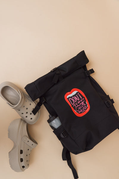 Cheer Roll Top Backpack in schwarz auf weißem Hintergrund. der Rucksack hat seitliche Gurt justierungen und Seitentaschen in einer ist eine Trinkflasch zu sehen. Auf dem Motiv ist eine weit offene Lippe mit Zähnen zu sehen. In der Öffnung vom Mund steht: Don't mess with my cheer squad please. Der Rucksack wurde auf beigem Hintergrund fotografiert. Neben dem Rucksack liegen Gartenlatschen mit Plateau.