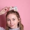 Junge Frau 20 Jahre alt, hat eine Halfbun Frisur mit einer bestickten Baumwollschleife auf dem Kopf, die sie mit der rechten Hand richtet. Sie ist im Halbportrait zu sehen, hat dunkelblonde Haare, braune Augen und trägt einen grauen Rollkragenpullover. Der Hintergrund ist rosa. Verkauft wird die Schleife/Haarspange.