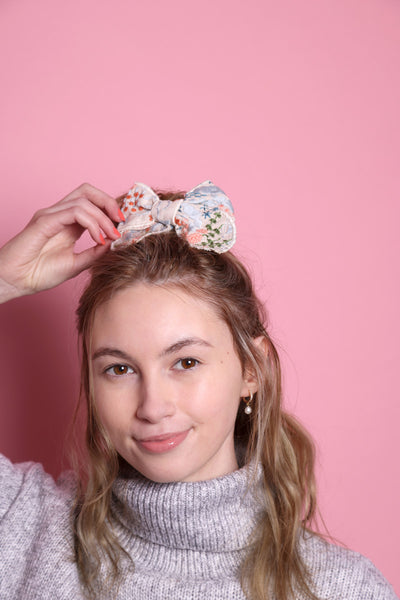 Junge Frau 20 Jahre alt, hat eine Halfbun Frisur mit einer bestickten Baumwollschleife auf dem Kopf, die sie mit der rechten Hand richtet. Sie ist im Halbportrait zu sehen, hat dunkelblonde Haare, braune Augen und trägt einen grauen Rollkragenpullover. Der Hintergrund ist rosa. Verkauft wird die Schleife/Haarspange.