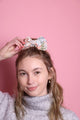 Junge Frau 20 Jahre alt, hat eine Halfbun Frisur mit einer bestickten Baumwollschleife auf dem Kopf, die sie mit der rechten Hand richtet. Sie ist im Halbportrait zu sehen, hat dunkelblonde Haare, braune Augen und trägt einen grauen Rollkragenpullover. Der Hintergrund ist rosa. Verkauft wird die Schleife/Haarspange.