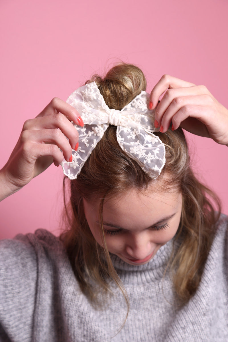 Junge Frau 20 Jahre alt, hat eine Halfbun Frisur mit einer offwhitefarbenen Schleife mit floralem Stickmuster auf dem Kopf, die sie mit den Händen richtet. Sie ist im Halbportrait, mit gesenktem Kopf zu sehen, hat dunkelblonde Haare, lächelt und trägt einen grauen Rollkragenpullover. Der Hintergrund ist rosa. Verkauft wird die Haarschleife als Haargummi.