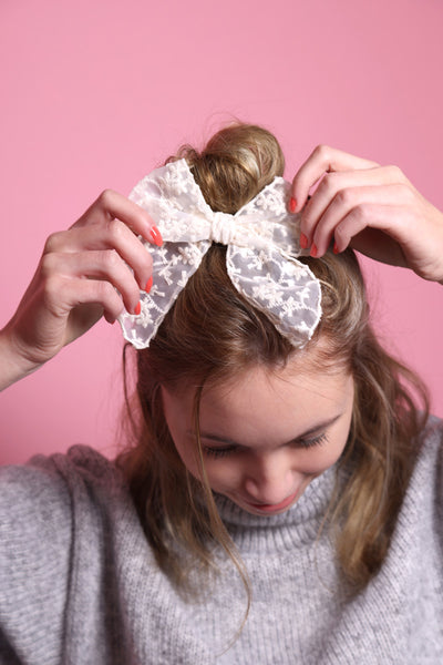 Junge Frau 20 Jahre alt, hat eine Halfbun Frisur mit einer offwhitefarbenen Schleife mit floralem Stickmuster auf dem Kopf, die sie mit den Händen richtet. Sie ist im Halbportrait, mit gesenktem Kopf zu sehen, hat dunkelblonde Haare, lächelt und trägt einen grauen Rollkragenpullover. Der Hintergrund ist rosa. Verkauft wird die Haarschleife als Haargummi.