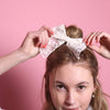 Junge Frau 20 Jahre alt, hat eine Halfbun Frisur mit einer weißen bestickten Schleife auf dem Kopf, die sie mit den Händen richtet. Sie ist im Halbportrait zu sehen,  hat dunkelblonde Haare, braune Augen und trägt einen grauen Rollkragenpullover. Sie lächelt und man sieht Perlenohrringe. Ihre Fingernägel sind Corallefarben. Der Hintergrund ist rosa. Verkauft wird die Schleife.