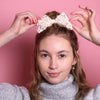 Junge Frau 20 Jahre alt, hat eine Halfbun Frisur mit einem weißer Haarschleife auf dem Kopf, die sie mit den Händen richtet. Sie ist im Halbportrait zu sehen, hat dunkelblonde Haare, braune Augen, lächelt und trägt einen grauen Rollkragenpullover. Ihr Nagellack ist Corallefarben. Der Hintergrund ist rosa. Verkauft wird die Haarschleife als Haargummi.