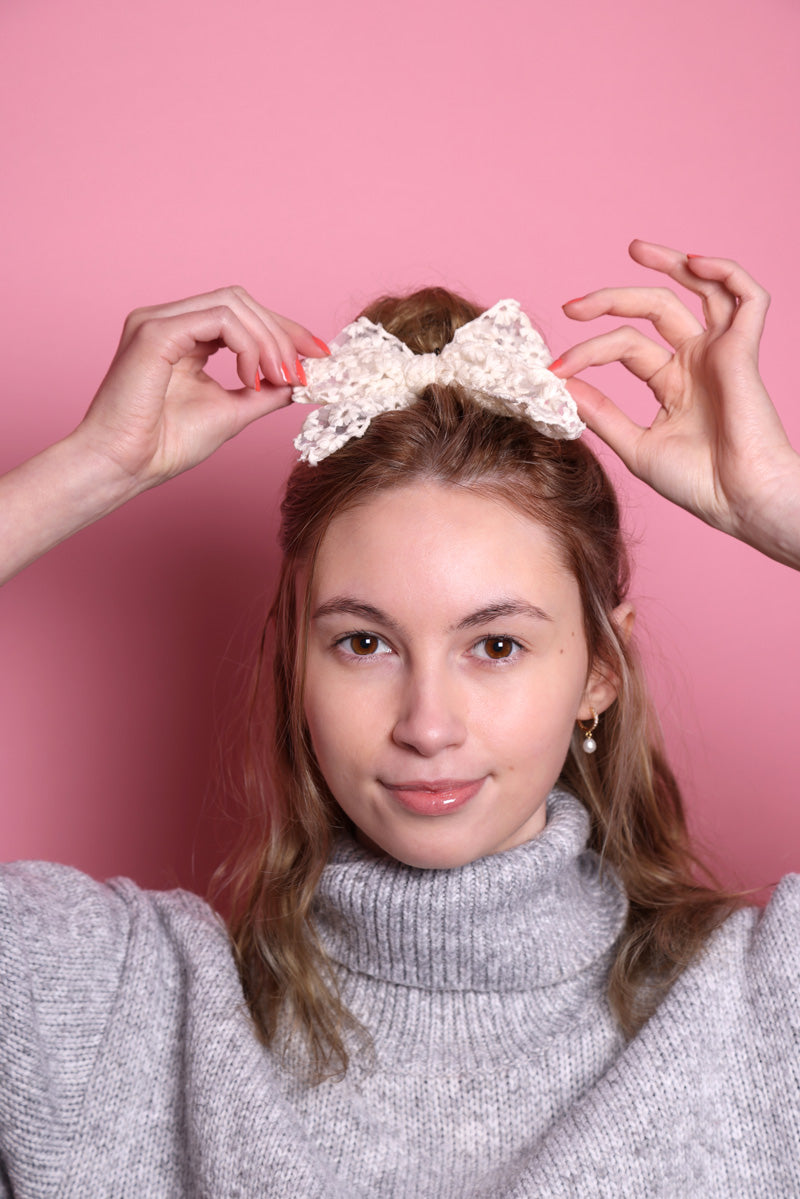 Junge Frau 20 Jahre alt, hat eine Halfbun Frisur mit einem weißer Haarschleife auf dem Kopf, die sie mit den Händen richtet. Sie ist im Halbportrait zu sehen, hat dunkelblonde Haare, braune Augen, lächelt und trägt einen grauen Rollkragenpullover. Ihr Nagellack ist Corallefarben. Der Hintergrund ist rosa. Verkauft wird die Haarschleife als Haargummi.