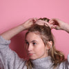Junge Frau 20 Jahre alt, hat eine Halfbun Frisur mit einem altrosanen Scrunchie auf dem Kopf, die sie mit den Händen richtet. Sie ist im Halbportrait zu sehen, leicht seitlich zu sehen, hat dunkelblonde Haare, braune Augen und trägt einen grauen Rollkragenpullover. Der Hintergrund ist rosa. Verkauft wird das Scunchie.