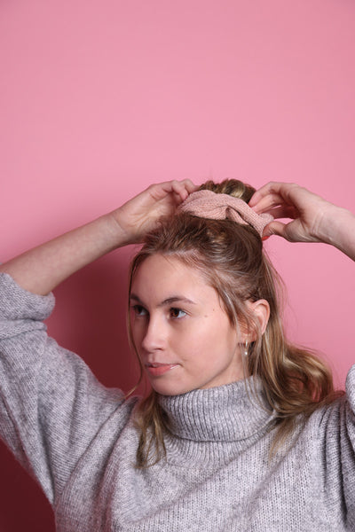 Junge Frau 20 Jahre alt, hat eine Halfbun Frisur mit einem altrosanen Scrunchie auf dem Kopf, die sie mit den Händen richtet. Sie ist im Halbportrait zu sehen, leicht seitlich zu sehen, hat dunkelblonde Haare, braune Augen und trägt einen grauen Rollkragenpullover. Der Hintergrund ist rosa. Verkauft wird das Scunchie.