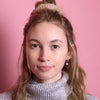 Junge Frau 20 Jahre alt, hat eine Halfbun Frisur mit einem cappuccino-farbenem Scrunchie auf dem Kopf. Sie ist im Halbportrait zu sehen, hat dunkelblonde Haare, braune Augen und trägt einen grauen Rollkragenpullover. Der Hintergrund ist rosa. Verkauft wird das Scunchie.