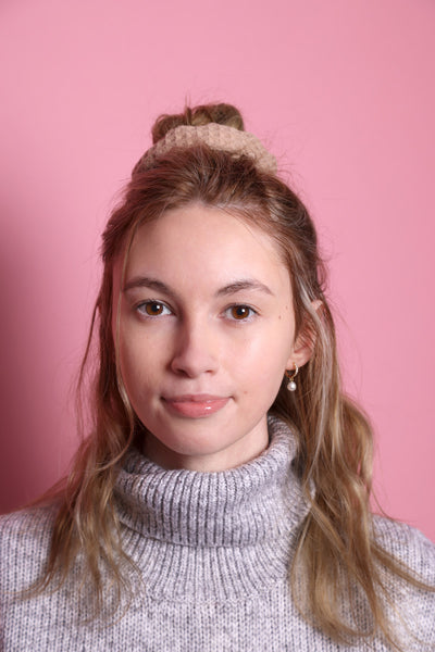 Junge Frau 20 Jahre alt, hat eine Halfbun Frisur mit einem cappuccino-farbenem Scrunchie auf dem Kopf. Sie ist im Halbportrait zu sehen, hat dunkelblonde Haare, braune Augen und trägt einen grauen Rollkragenpullover. Der Hintergrund ist rosa. Verkauft wird das Scunchie.