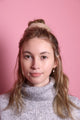Junge Frau 20 Jahre alt, hat eine Halfbun Frisur mit einem cappuccino-farbenem Scrunchie auf dem Kopf. Sie ist im Halbportrait zu sehen, hat dunkelblonde Haare, braune Augen und trägt einen grauen Rollkragenpullover. Der Hintergrund ist rosa. Verkauft wird das Scunchie.