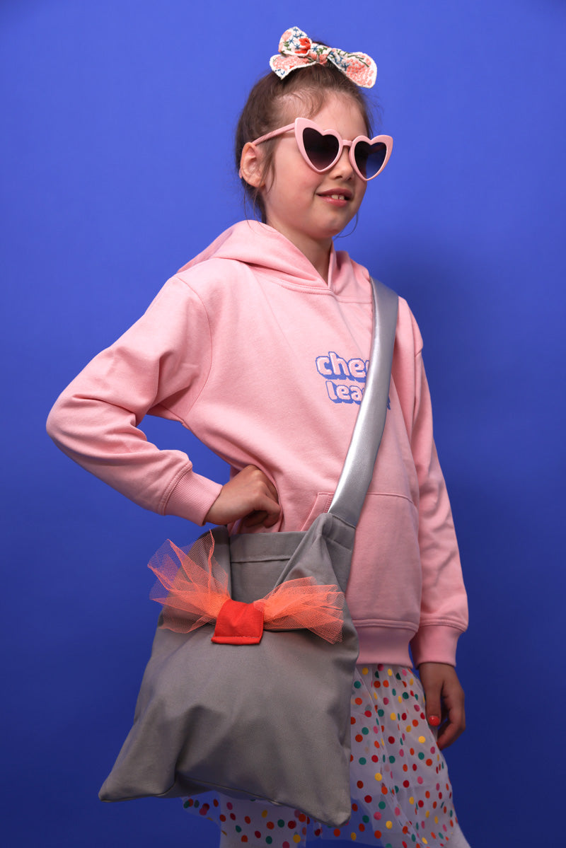 Ein kleines Mädchen mit mittelblonden Haaren und einer bestickten Schleife auf dem Kopf steht vor einer blauen Wand. Sie trägt zudem eine rosafarbene, abgedunkelte Herzsonnenbrille, einen gepunkteten Rock, eine graue Tasche mit silbernem Henkel und Corallefabener Schleife. Außerdem hat sie einen rosafarbenen Hoodie mit Kangerootasche und Kaputze an. Auf dem Hoodie steht Cheerleader. Sie steht seitlich und hat einen Arm in der Hüfte abgelehnt und lächelt freundlich.