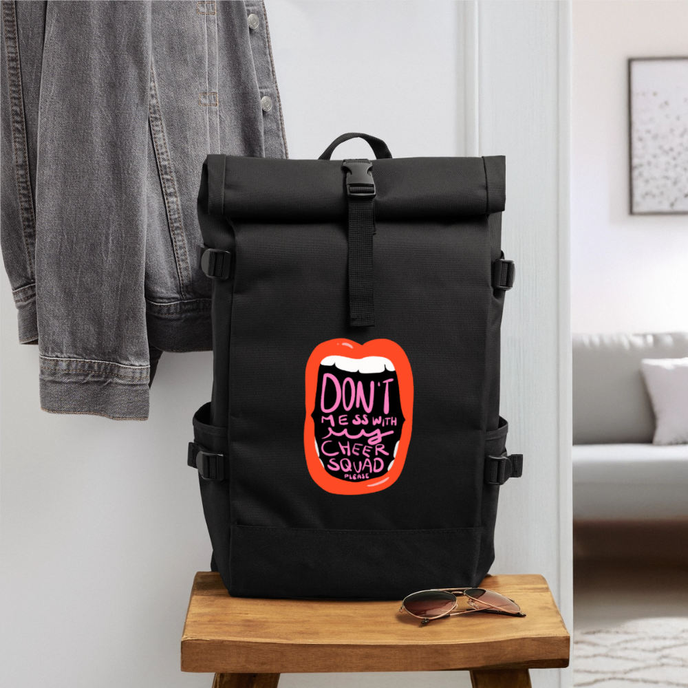 Cheer Roll Top Backpack in schwarz. Der Rucksack hat seitliche Gurt-Justierungen und Seitentaschen für z.B. Flaschen. Auf dem Motiv ist eine weit offene Lippe mit Zähnen zu sehen. In der Öffnung vom Mund steht: Don't mess with my cheer squad please. Der Rucksack wurde in einem Flur auf einem Holz-Hocker mit Jeansjacke dahinter  und Sonnenbrille davor fotografiert. Rechts sieht man den Durchgang in ein helles Wohnzimmer mit Couch, Bild, und Teppich.