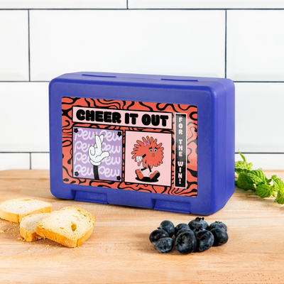 Cheerleader Lunchbox in royal blue steht vor weißem Hintergrund auf weißem Untergrund. Auf der Dose ist ein Comicmitiv mit der Aufschrift: Cheer it out for the Win auf einem rot schwarzem Muster. Außerdem sieht man hier einen Pompom mit Armen und Meinen und Haarschleife, wie auch Augen. Die Pompomfigur pfeift. Zudem gibt es noch eine Comichand mit gekreuzten Fingern. Die Dose steht auf einem Holzhintergrund vor einer weißen Fliesenwand. Auf der Holzarbeitsfläche sind Zwieback, ein paar Blaubeeren und Minze.