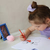 Ein kleines Peeweemädchen sitzt an einem weißen Schreibtisch vor einer weißen Wand und malt in einem Cheerleading Ausmalbuch ein Motiv mit einer Schleife aus. Sie benutzt dafür einen roten Stift. Neben ihr liegt ein rosa Herzplüsch-Stift und es steht ein blauer Rahmen mit einer Cheerpostkarte. SIe trägt ein helllilanes Scrunchie und einen lilafarbenen Pullunder. Ihre Nägel sind Pink.