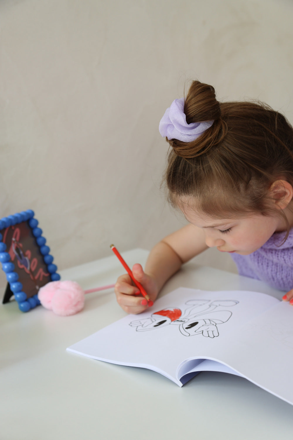 Ein kleines Peeweemädchen sitzt an einem weißen Schreibtisch vor einer weißen Wand und malt in einem Cheerleading Ausmalbuch ein Motiv mit einer Schleife aus. Sie benutzt dafür einen roten Stift. Neben ihr liegt ein rosa Herzplüsch-Stift und es steht ein blauer Rahmen mit einer Cheerpostkarte. SIe trägt ein helllilanes Scrunchie und einen lilafarbenen Pullunder. Ihre Nägel sind Pink.