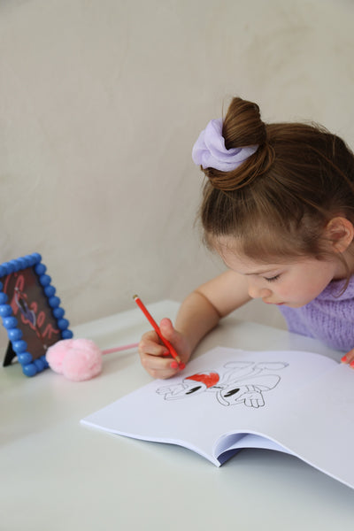 Ein kleines Peeweemädchen sitzt an einem weißen Schreibtisch vor einer weißen Wand und malt in einem Cheerleading Ausmalbuch ein Motiv mit einer Schleife aus. Sie benutzt dafür einen roten Stift. Neben ihr liegt ein rosa Herzplüsch-Stift und es steht ein blauer Rahmen mit einer Cheerpostkarte. SIe trägt ein helllilanes Scrunchie und einen lilafarbenen Pullunder. Ihre Nägel sind Pink.