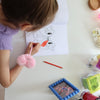 Ein kleines Peeweemädchen sitzt an einem weißen Schreibtisch vor einer weißen Wand und malt in einem Cheerleading Ausmalbuch ein Motiv mit einer Schleife aus. Sie benutzt dafür einen roten Stift. Neben ihr liegt ein rosa Herzplüsch-Stift und es steht ein blauer Rahmen mit einer Cheerpostkarte. SIe trägt ein helllilanes Scrunchie und einen lilafarbenen Pullunder. Ihre Nägel sind Pink. Man sieht zu dem noch den Malkasten, eine gelbe Vase, leuchtend bunte Organizer-dosen und Stiftehalter.