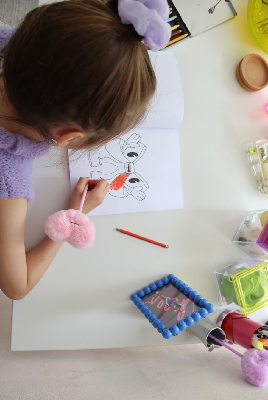 Ein kleines Peeweemädchen sitzt an einem weißen Schreibtisch vor einer weißen Wand und malt in einem Cheerleading Ausmalbuch ein Motiv mit einer Schleife aus. Sie benutzt dafür einen roten Stift. Neben ihr liegt ein rosa Herzplüsch-Stift und es steht ein blauer Rahmen mit einer Cheerpostkarte. SIe trägt ein helllilanes Scrunchie und einen lilafarbenen Pullunder. Ihre Nägel sind Pink. Man sieht zu dem noch den Malkasten, eine gelbe Vase, leuchtend bunte Organizer-dosen und Stiftehalter.