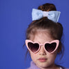Ein kleines Mädchen mit lilafarbenem Wollpullunder steht vor einer blauen Wand. Sie trägt eine rosafarbene Herzbrille mit dunklen Gläsern. Ihre mittelblonden Haare sind im Dutt und sie trägt eine hellblaue gepolsterte Schleife mit Vintage Spitze drum. 