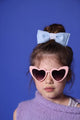 Ein kleines Mädchen mit lilafarbenem Wollpullunder steht vor einer blauen Wand. Sie trägt eine rosafarbene Herzbrille mit dunklen Gläsern. Ihre mittelblonden Haare sind im Dutt und sie trägt eine hellblaue gepolsterte Schleife mit Vintage Spitze drum. 